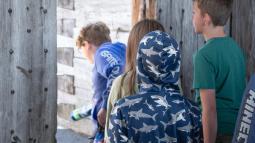 Children in colorful clothing climb through a wooden door with large bolts on it.