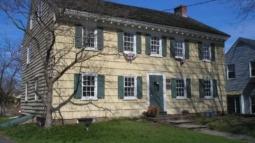 A yellow colonial‑style house with green shutters and a steep dark roof stands behind a leafless tree, with a stone walkway leading to its green front door.