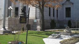 A light‑stone courthouse stands behind a grassy area with a tree, a small cannon monument, and a historical marker noting the site of an early borough courthouse built before the Revolution.