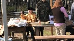 Person wearing colonial clothing sitting at a wooden table with a mannequin on the table.