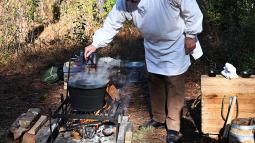 Man in a white smock bending over a campfire dipping a candle
