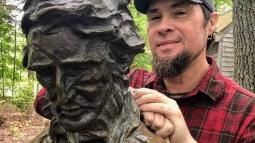 A man in a plaid shirt leans on a bronze monument of Henry David Thoreau