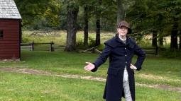A man in 18th century period clothing stands in front of a campfire pit.