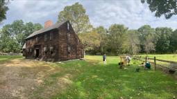 A historic wooden house in a field with people dressed in colonial clothing outside, and a cider pre
