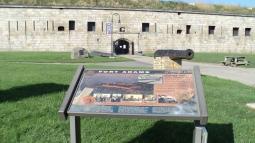 A stone fort wall with an arched entrance rises behind a grassy area where an informational sign, an old cannon, and a picnic table sit under a bright blue sky.