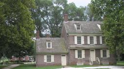 A brick colonial‑style house with cream shutters and multiple chimneys stands amid green grass and tall trees.
