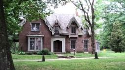 A large brick Gothic‑style house with steep gables and tall narrow windows stands among mature trees on a grassy lawn.
