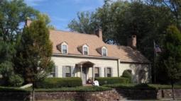 A tan colonial‑style house with dormer windows, brick chimneys, and a columned porch stands behind trimmed hedges and a stone retaining wall, with an American flag flying beside it.