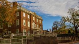 A large brick building with white trim and classical details stands behind a tall white fence, framed by autumn trees under a partly cloudy sky.