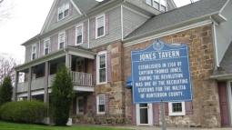 A stone‑and‑shingle historic building with a columned porch stands behind a blue marker identifying it as Jones Tavern, established in 1760.