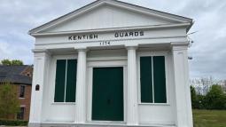 A small white historic building with four columns and green shutters stands at a roadside; the sign above the door reads “Kentish Guards 1774.”