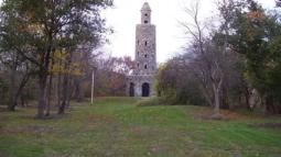 A tall cylindrical stone tower with small windows and an arched entrance stands in a grassy clearing surrounded by trees showing early autumn color.