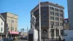 A colonial era militia statue enclosed by a circular black fence stands in a small plaza with an American flag behind it, surrounded by multi‑story city buildings under a bright blue sky.