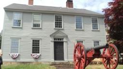 Colonial era 2-story manor house with 9 windows trimmed in white, with 2 chimneys and a red cannon in the front yard.