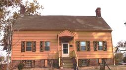 A two‑story yellow colonial house with dark brown trim and twin brick chimneys stands behind a small front porch and brick walkway.