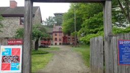 A wooden entrance gate stands at the Old Barracks Museum, built from two tall posts and a horizontal beam.  Beyond the gate, a gravel path leads to historic stone and brick buildings with red trim, surrounded by trees and greenery.