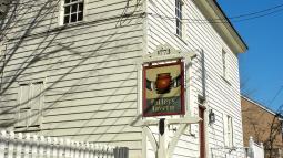 A white historic building with a stone foundation and an exterior stairway stands behind a sign reading “Potter’s Tavern 1773” topped with a brown pot illustration.