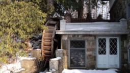 A rustic stone mill with a large wooden water wheel stands in the snow among evergreen trees.
