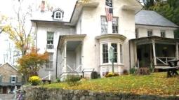 A large light‑colored house with multiple gables, a stone wall in front, and an American flag above the porch sits among autumn trees with fallen leaves.