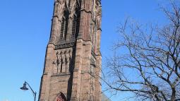 A tall brown stone Gothic‑style church tower with pointed arches and ornate spires rises above trees under a bright blue sky.
