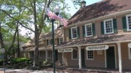 A tan historic building with green shutters and a columned porch stands along a brick sidewalk, shaded by large trees and marked by a streetlamp flying an American flag.