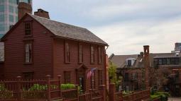 A reddish‑brown historic wooden house with tall narrow windows and an American flag at the entrance stands behind a matching rod iron fence.
