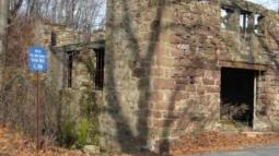 The stone ruins of an old grist mill stand among leafless trees, with a blue historical marker identifying the site beside the weathered walls.