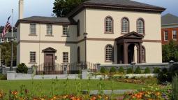 A beige building with brown trim and a columned portico stands behind a garden of bright flowers and neatly trimmed shrubs, bordered by a wrought‑iron fence and stone posts.