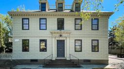 A light‑colored historic building with dark shutters and a central double‑door entrance sits on a corner lot, featuring three dormer windows on the roof and a short staircase leading up from the sidewalk.