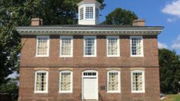 A two‑story brick Federal style building with white‑trimmed windows and a central cupola stands on a grassy lawn beneath a bright blue sky.