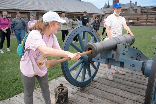 A young woman is using a long wooden q-tip like stick to clean a cannon while her friends watch on.