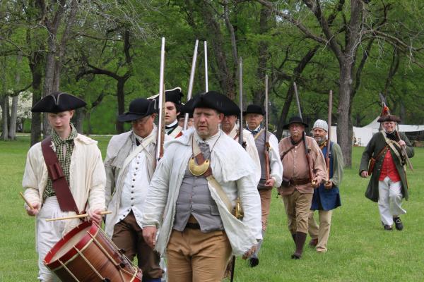 Men in colonial clothing marching behind a drummer.