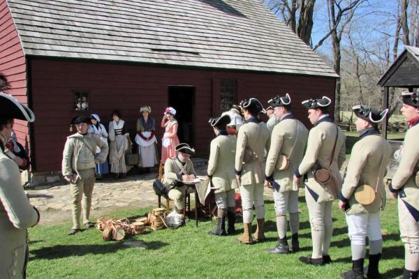 Men in grey Continental soldier uniforms stand in line near a large wooden building.