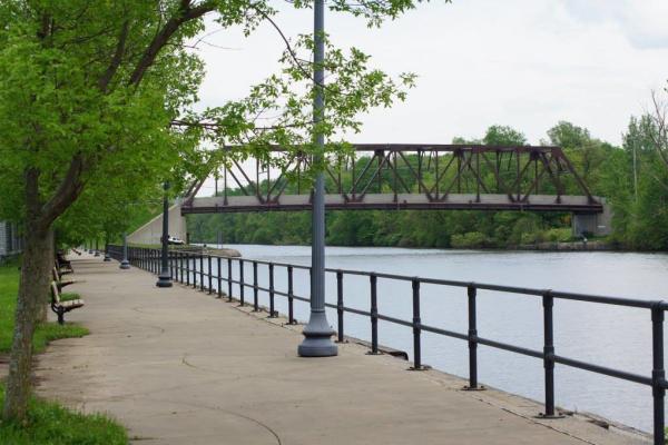 Along a sidewalk, you can see a metal railing and just beyond that, a river and a metal bridge.