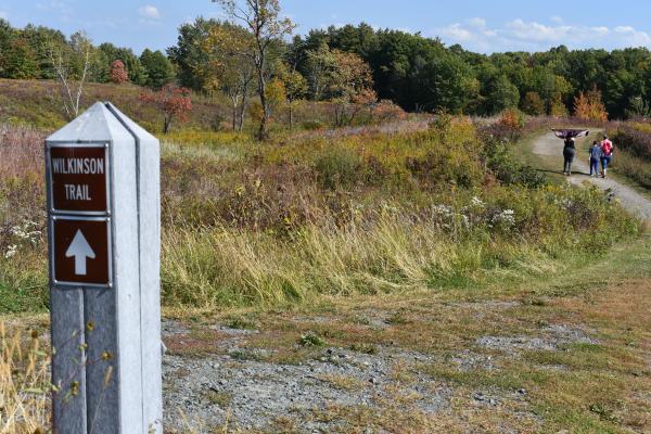 Wilkinson Trail marker in foreground, trail with 3 hikers cutting through meadow