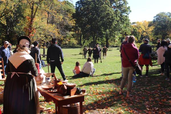 People in colonial clothing mingle with modern visitors in a field with trees in autumn color