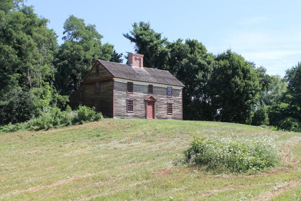 A two story wooden colonial house sits on a hill surrounded by green trees.