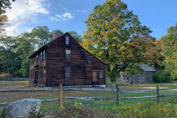 A wooden colonial era house stands in a grassy field surrounded by trees and a wooden rail fence