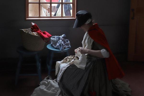 A women in 1775 clothing sits near a window in the Smith House wrapping bread with a cloth