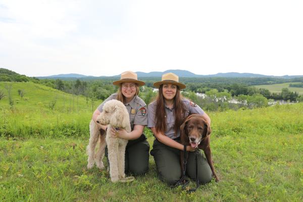 Two women in park ranger uniforms kneel next to two dogs on leash.