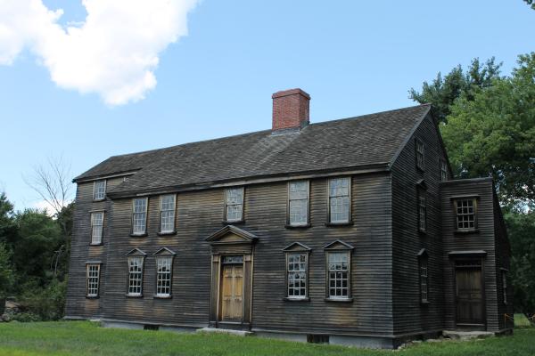 A three story colonial saltbox style house made of wood. It is surrounded by green grass and trees.