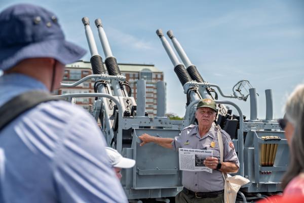 A ranger stands in front of a gun mount speaking to members of the public.