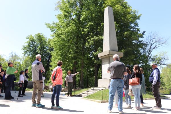 A park ranger stands in front of a towering stone monument pointing as visitors stand around.