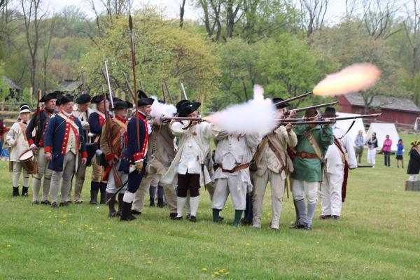 A line of Revolutionary War re-enactors in period uniforms fire muskets during a demonstration at Jockey Hollow. Smoke and muzzle flashes rise as the volley is discharged. Behind them, more re-enactors stand at attention with fixed bayonets.