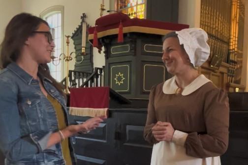 One woman in modern clothes gestures in conversation to another women dressed in historical clothing as Abigail Adams. They are standing inside historic St. John's Church. 