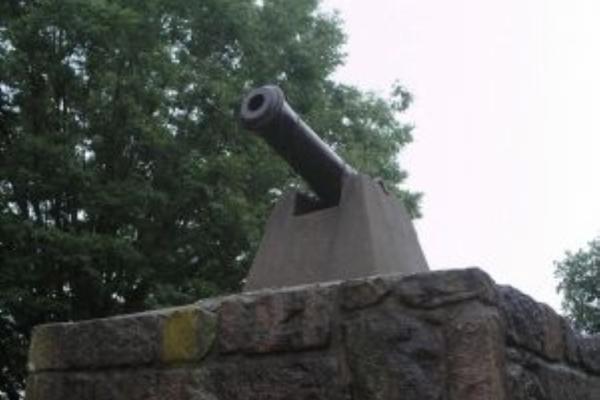 A black cannon sits atop a small stone structure with a glass window, surrounded by green trees under an overcast sky.