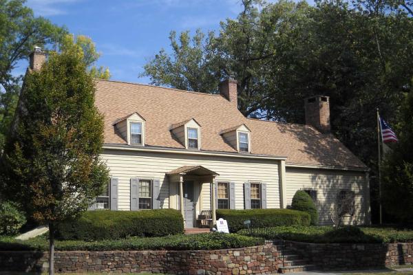 A tan colonial‑style house with dormer windows, brick chimneys, and a columned porch stands behind trimmed hedges and a stone retaining wall, with an American flag flying beside it.