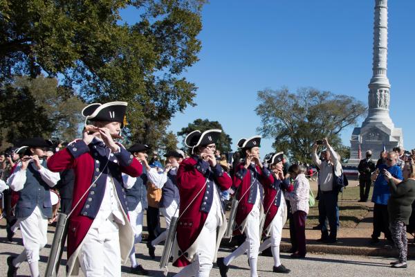 Fifes and drum wearing colonial uniforms with red jackets, march by the Yorktown Victory monument