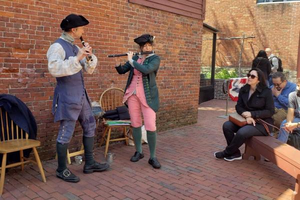 Two people wearing colonial garb and playing instruments while a few people watch