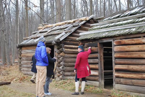 Two men in colonial living history clothing standing in front of log cabins.